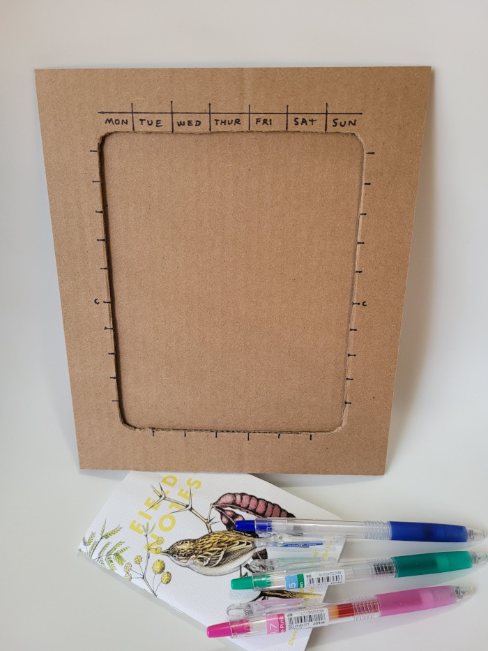 Cardboard template with markings for all of the calendar grid lines. Shown above notebook and pens.