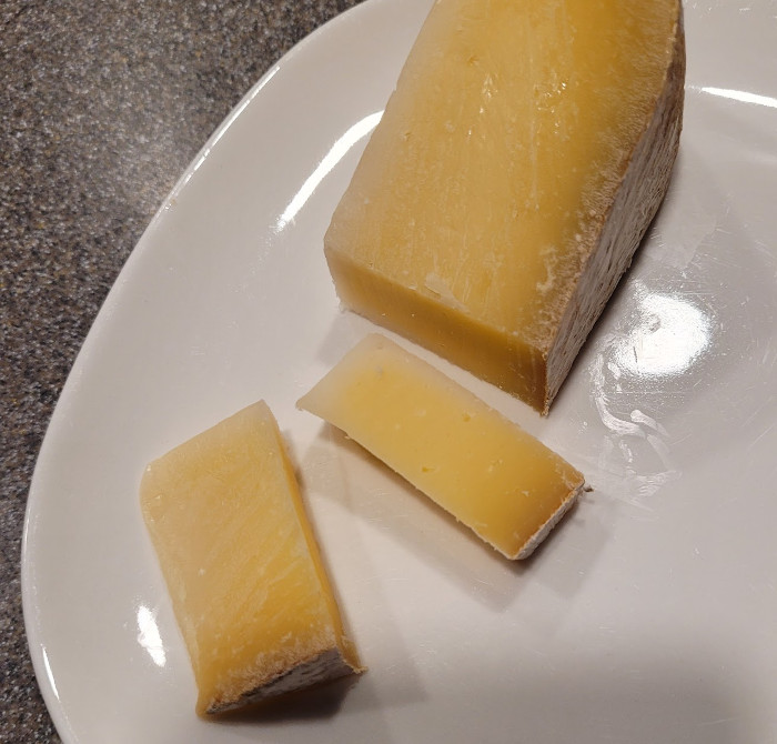 Photo of a wedge of the cheese with two slices. It’s mostly pale yellow on the inside and dusty gray on the outside.